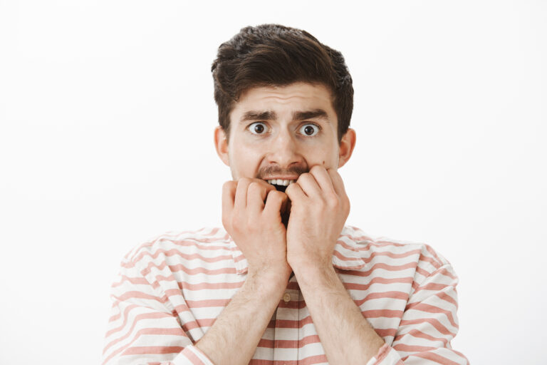 Studio shot of scared trembling poor european guy in stripes shirt, biting fingers and staring afraid at camera, being frightened to death, looking scary movie alone in dark, standing over gray wall